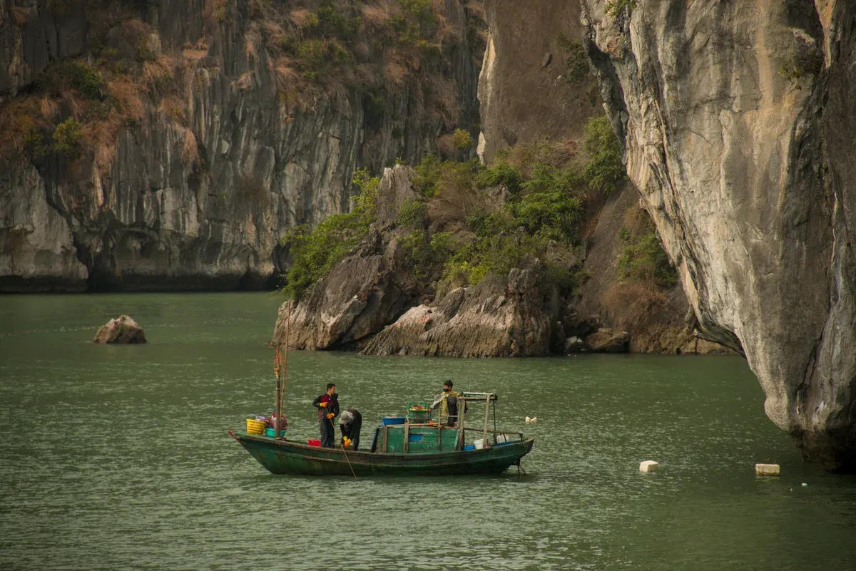 Fishermen in a small boat below a limestone cliff in Bai Tu Long Bay, Halong Bay cruise